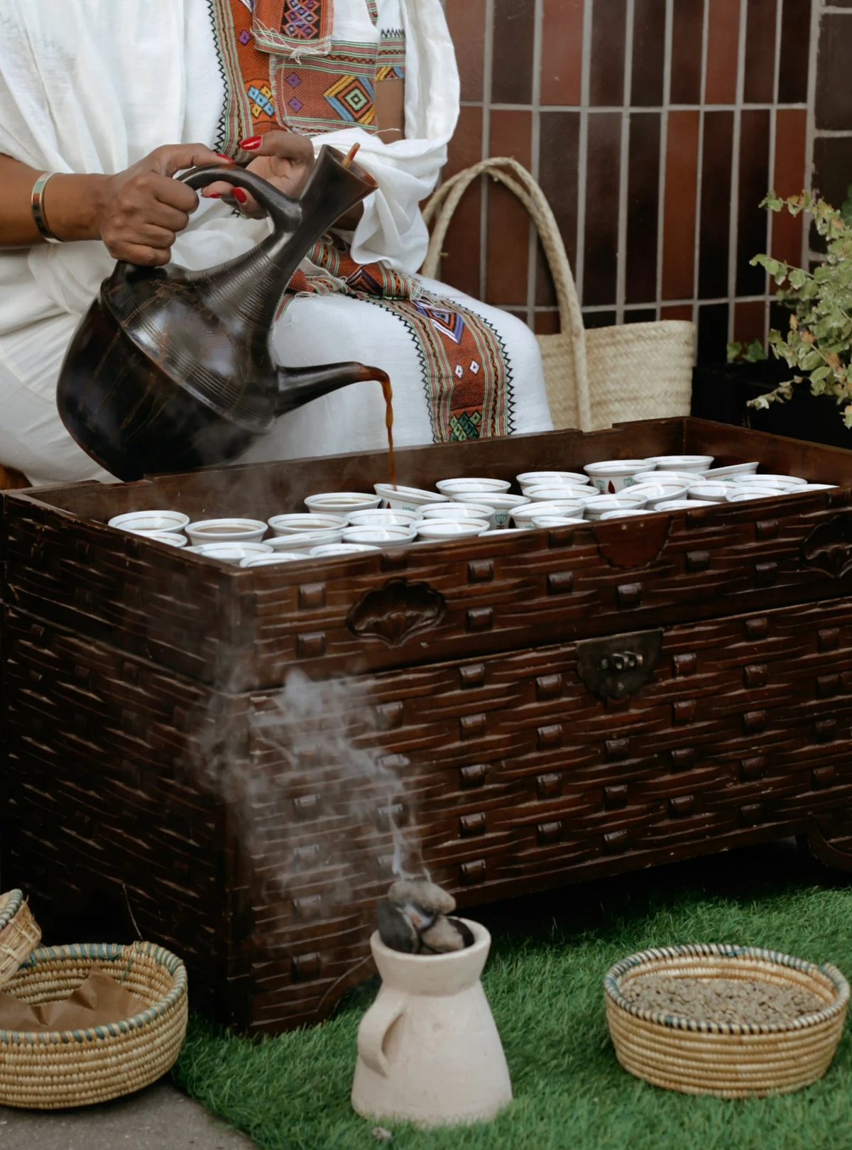 A jebena pouring coffee into rows of small cups beside rising incense